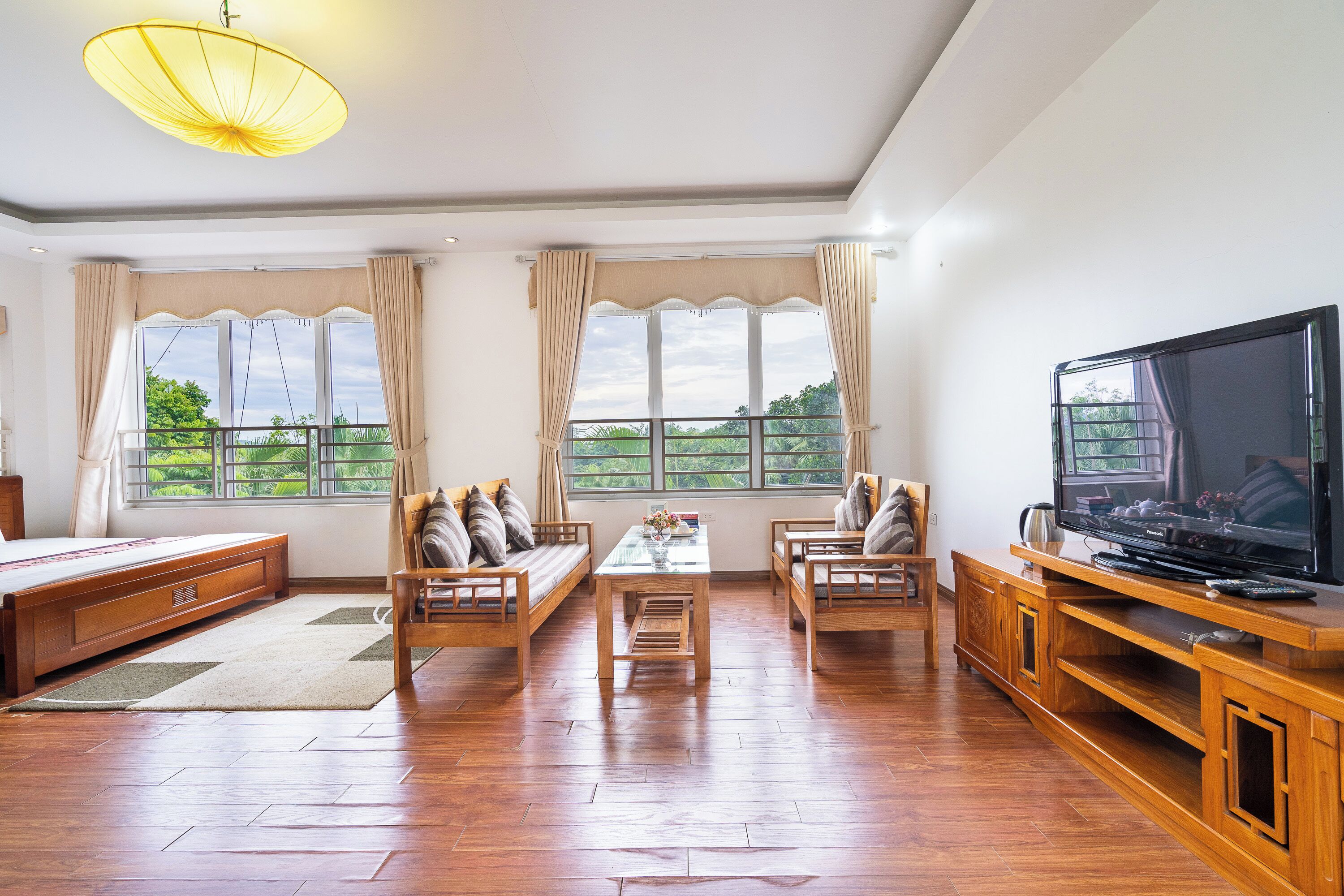 A spacious living room with a wooden floor, a sofa set, a TV on a wooden stand, and large windows with curtains.