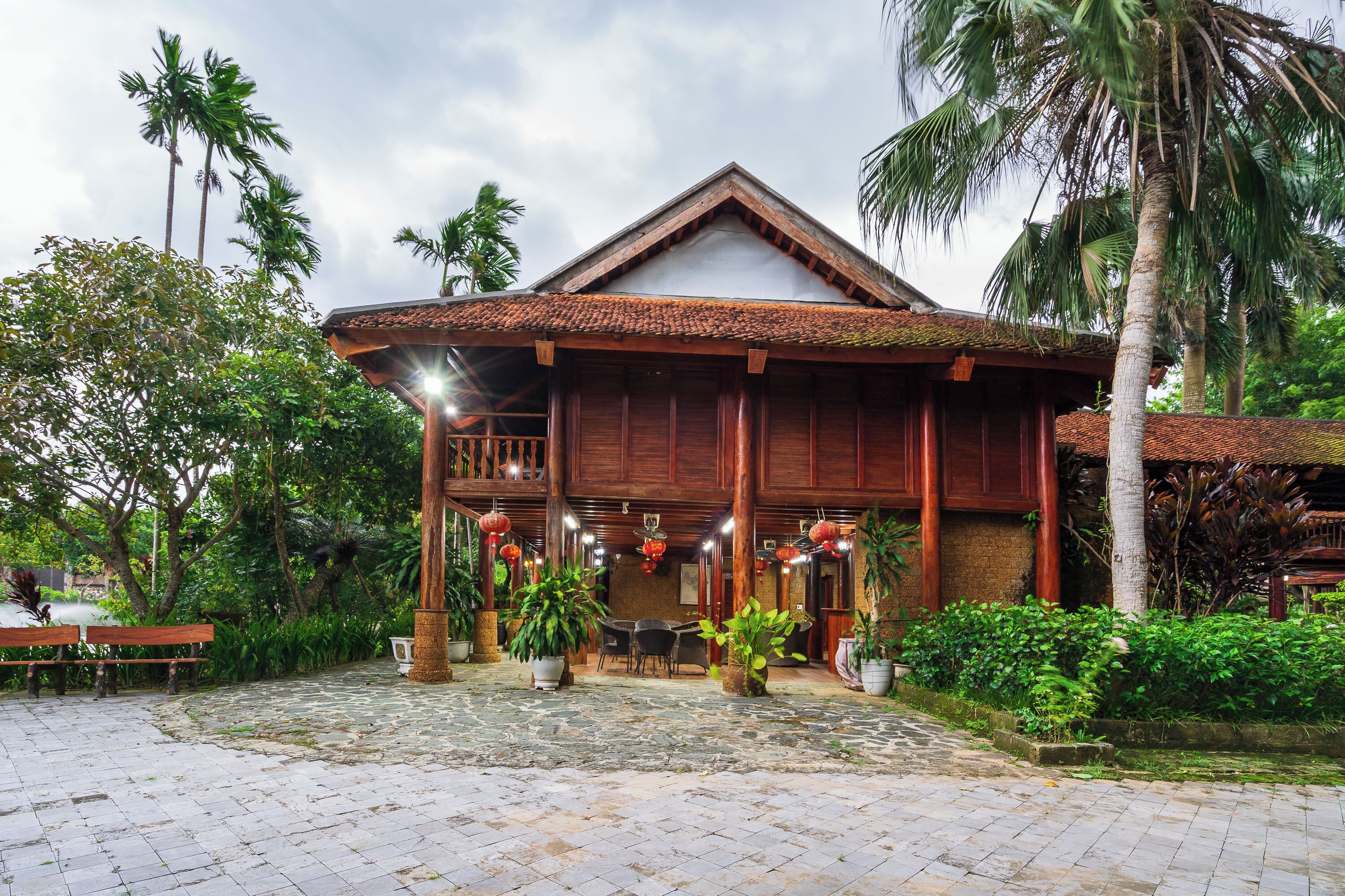A traditional wooden building with a tiled roof, surrounded by lush greenery and palm trees.