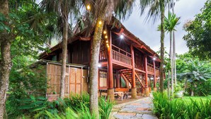 A wooden house with a balcony, surrounded by lush greenery and a stone pathway.