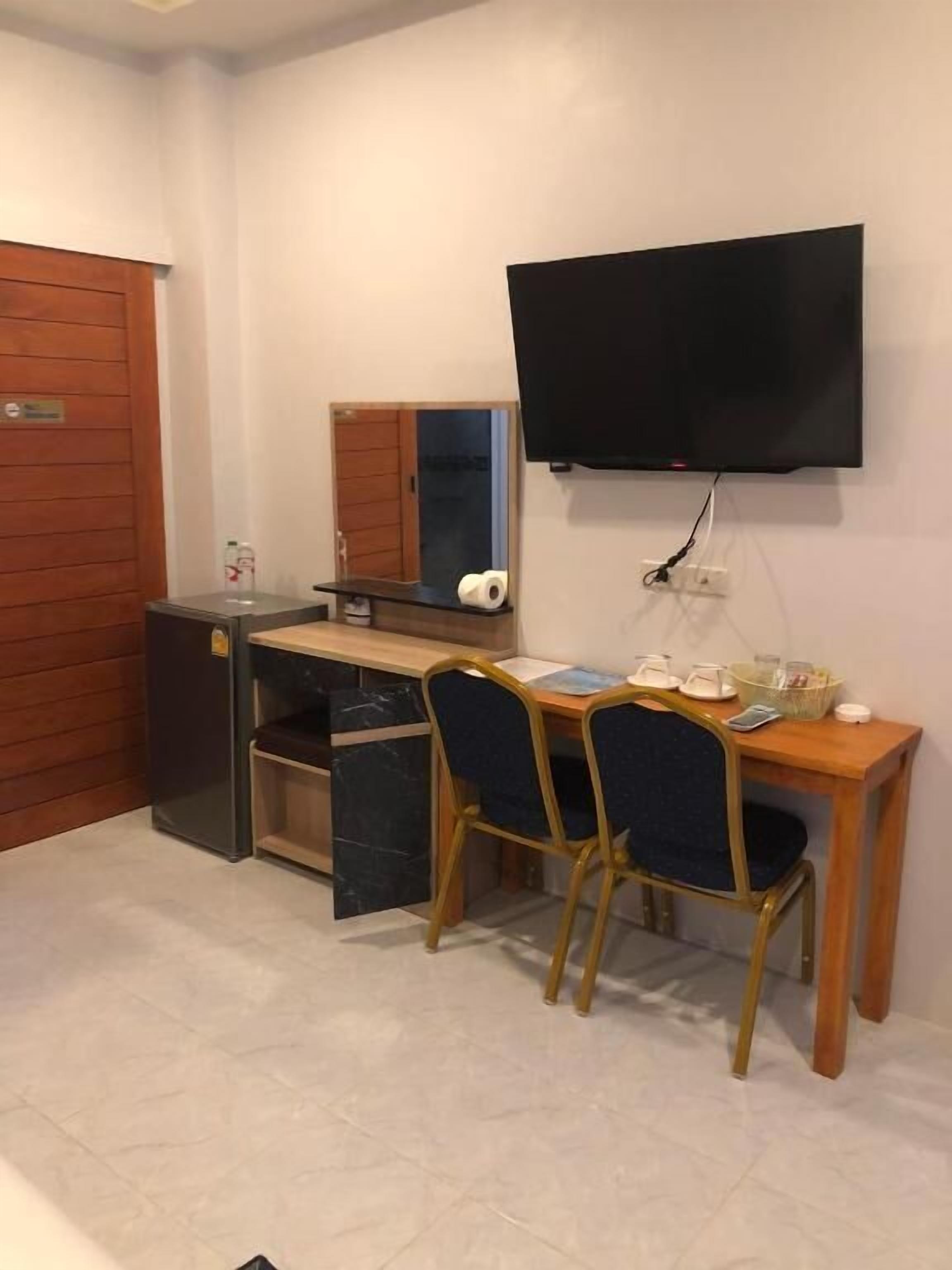 A compact room with a wooden desk, two chairs, a flat-screen TV mounted on the wall, a small cabinet, and a door.