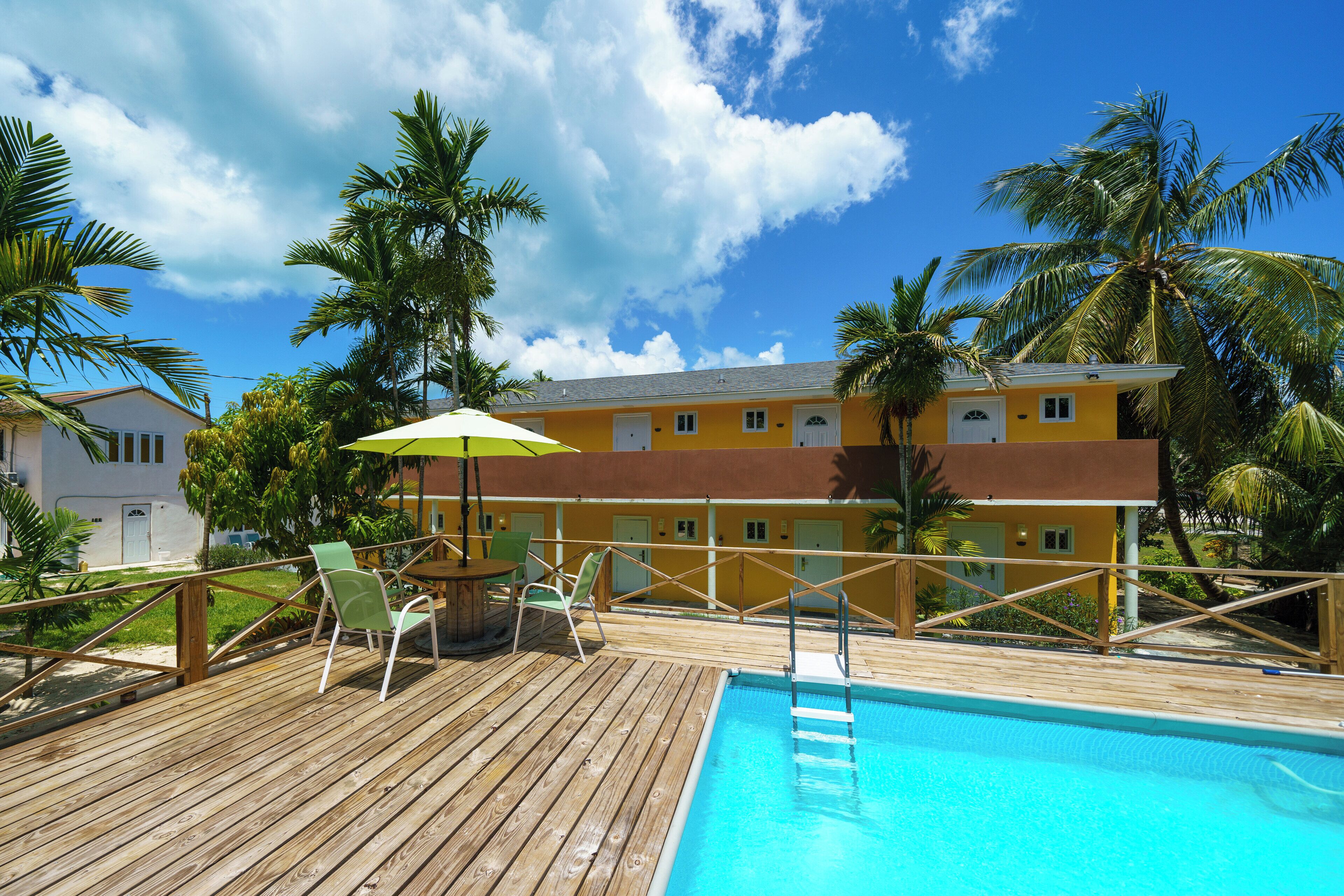 A wooden deck with a pool, outdoor chairs, and a yellow building with a brown roof.