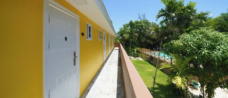 A yellow building with a white door, a concrete pathway, and a pool area with palm trees.
