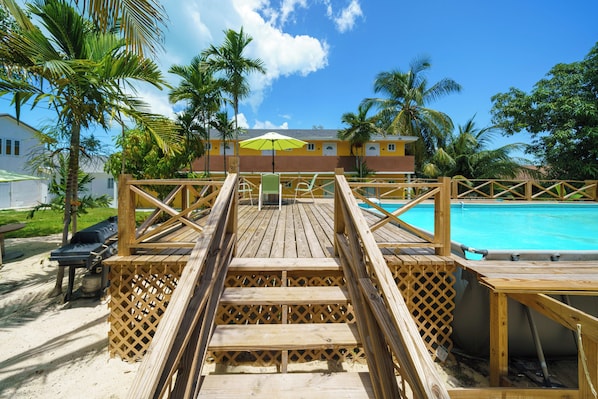 A wooden deck leading to a pool area with a view of a building in the background. - Hilton's Haven (Tarpum Bay)