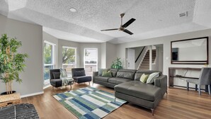 A spacious living room with a large sectional sofa, a ceiling fan, and a view of the outdoors through large windows.