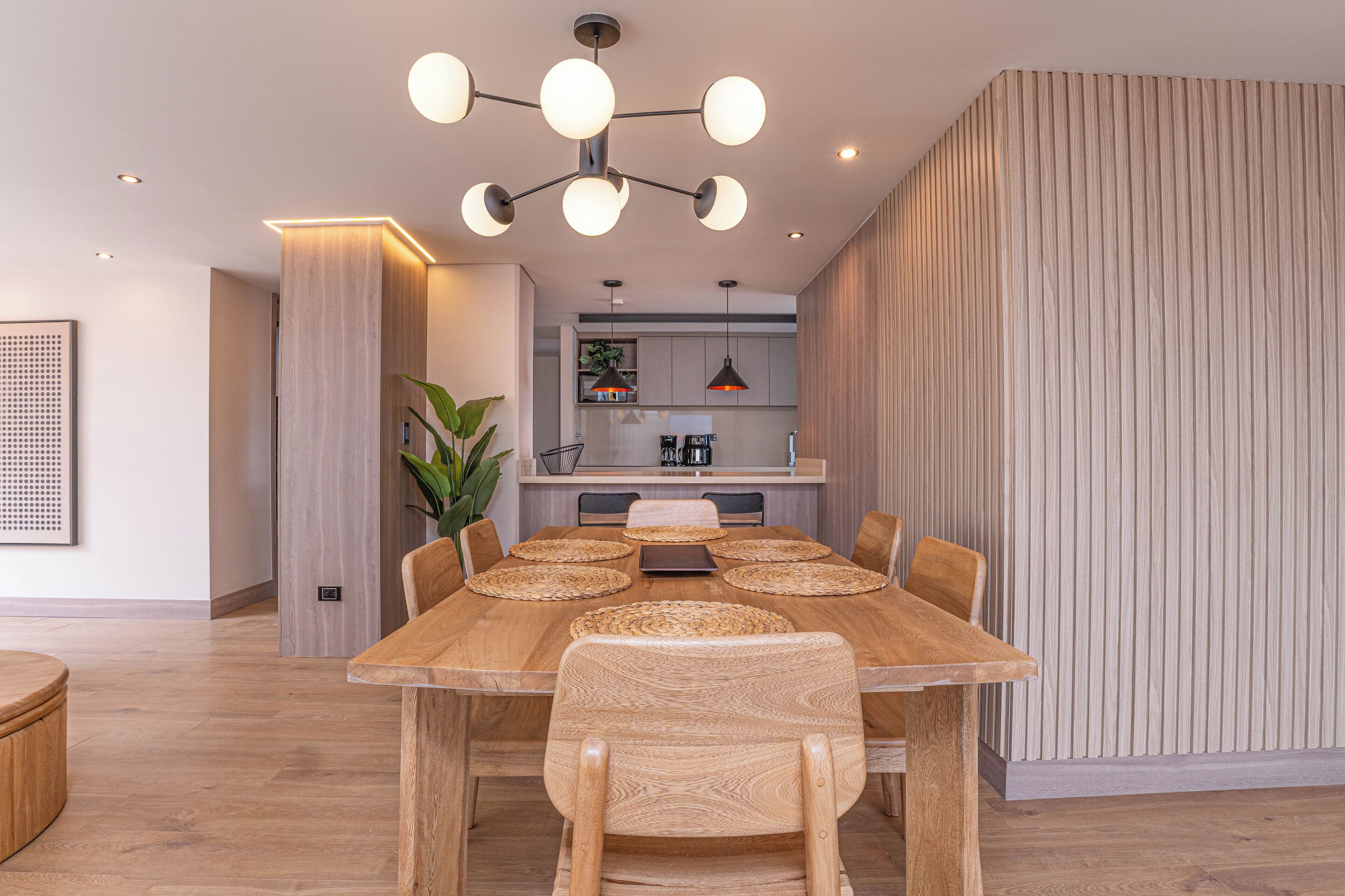 A modern dining area with a wooden table and chairs, a pendant light, and a kitchen in the background.