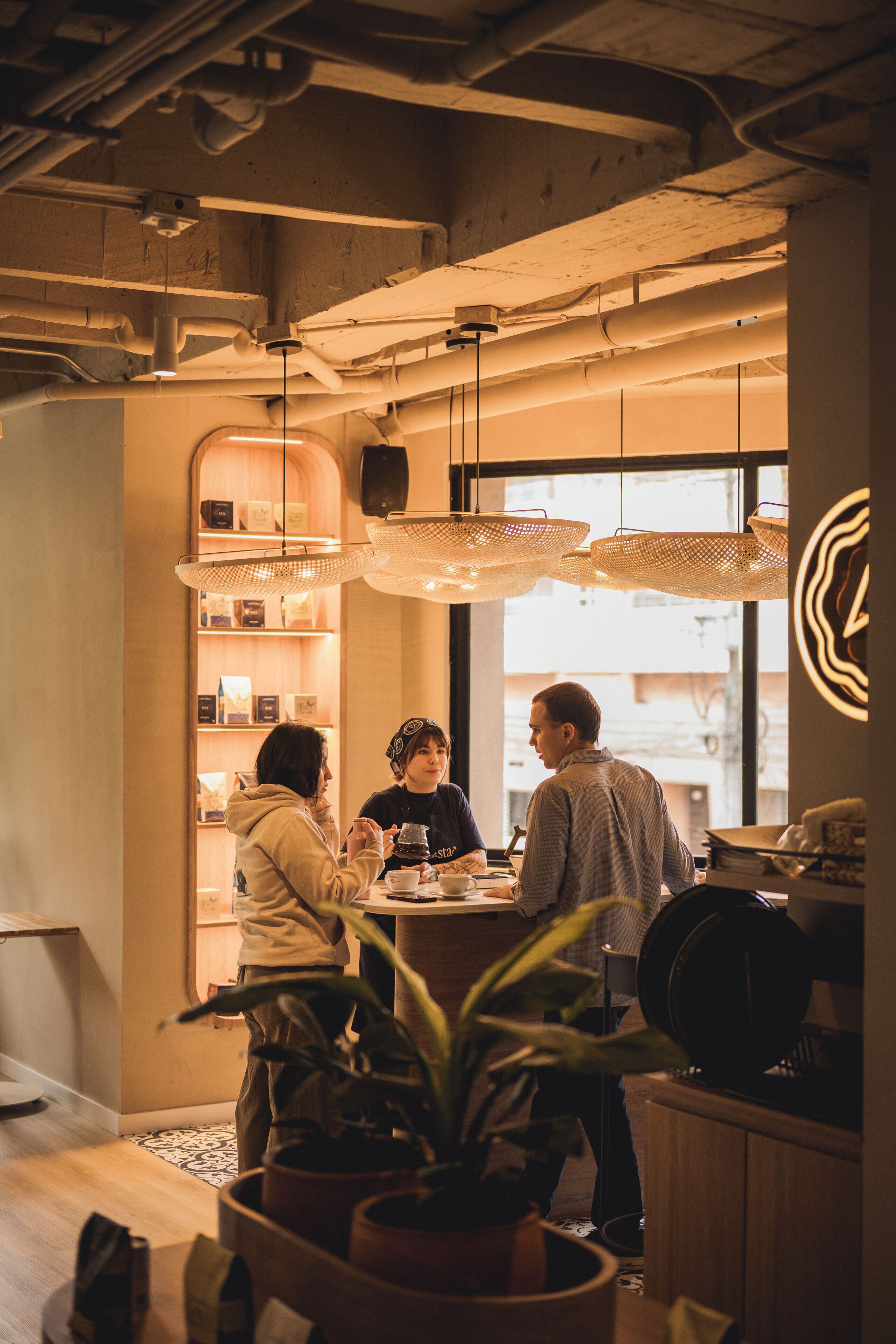 Een interieur met zichtbare leidingen, hangende geweven lampen en een houten toonbank met een koffieapparaat.