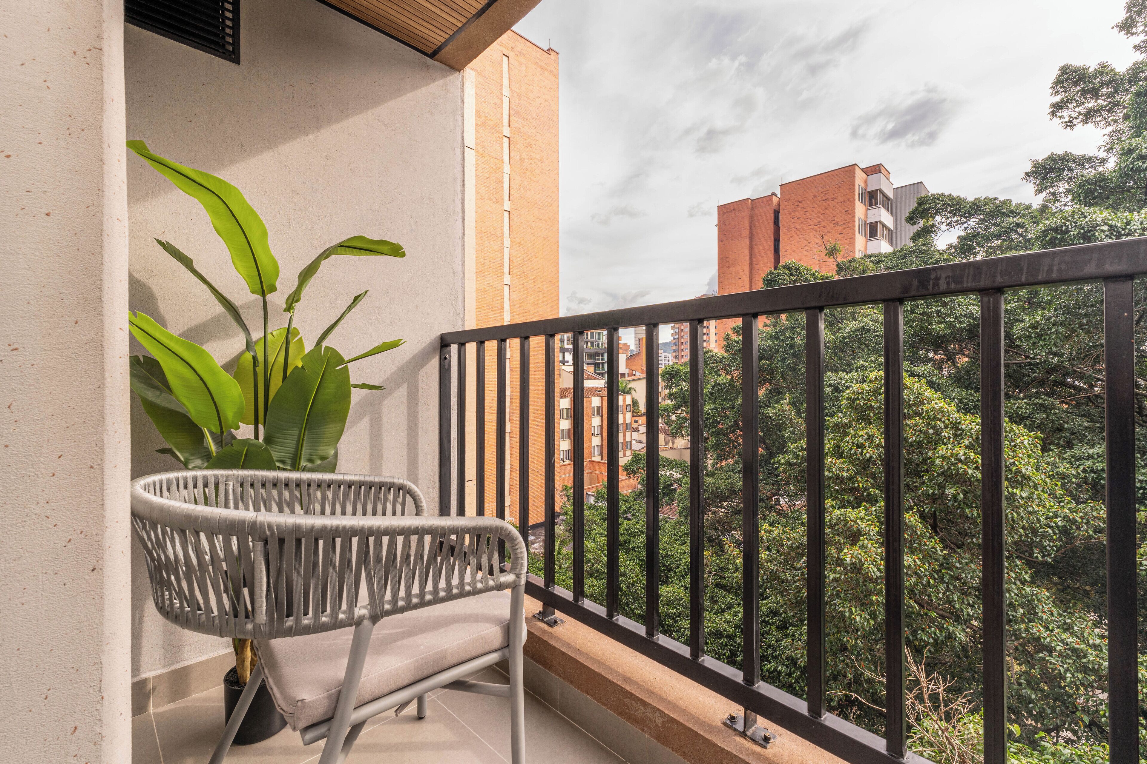 Ein Balkon mit einem weißen Korbstuhl, einem Blumentopf und Blick auf eine Stadtlandschaft mit Gebäuden und Bäumen.