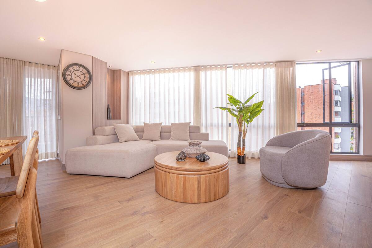 Un salon moderne avec un canapé d’angle, une table basse ronde en bois, un fauteuil gris et une grande fenêtre avec des rideaux.