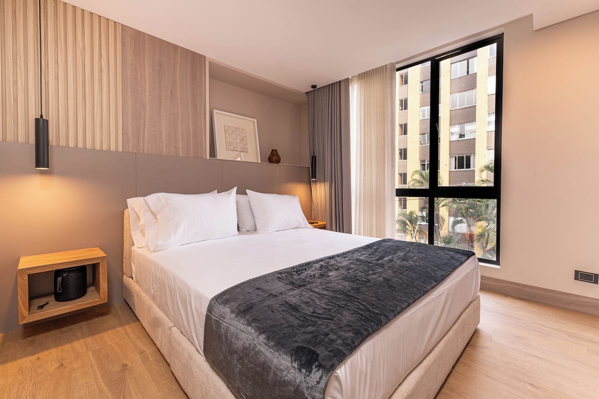 Une chambre d’hôtel moderne avec un grand lit, une table de chevet en bois, un tableau encadré et une grande fenêtre donnant sur des immeubles.
