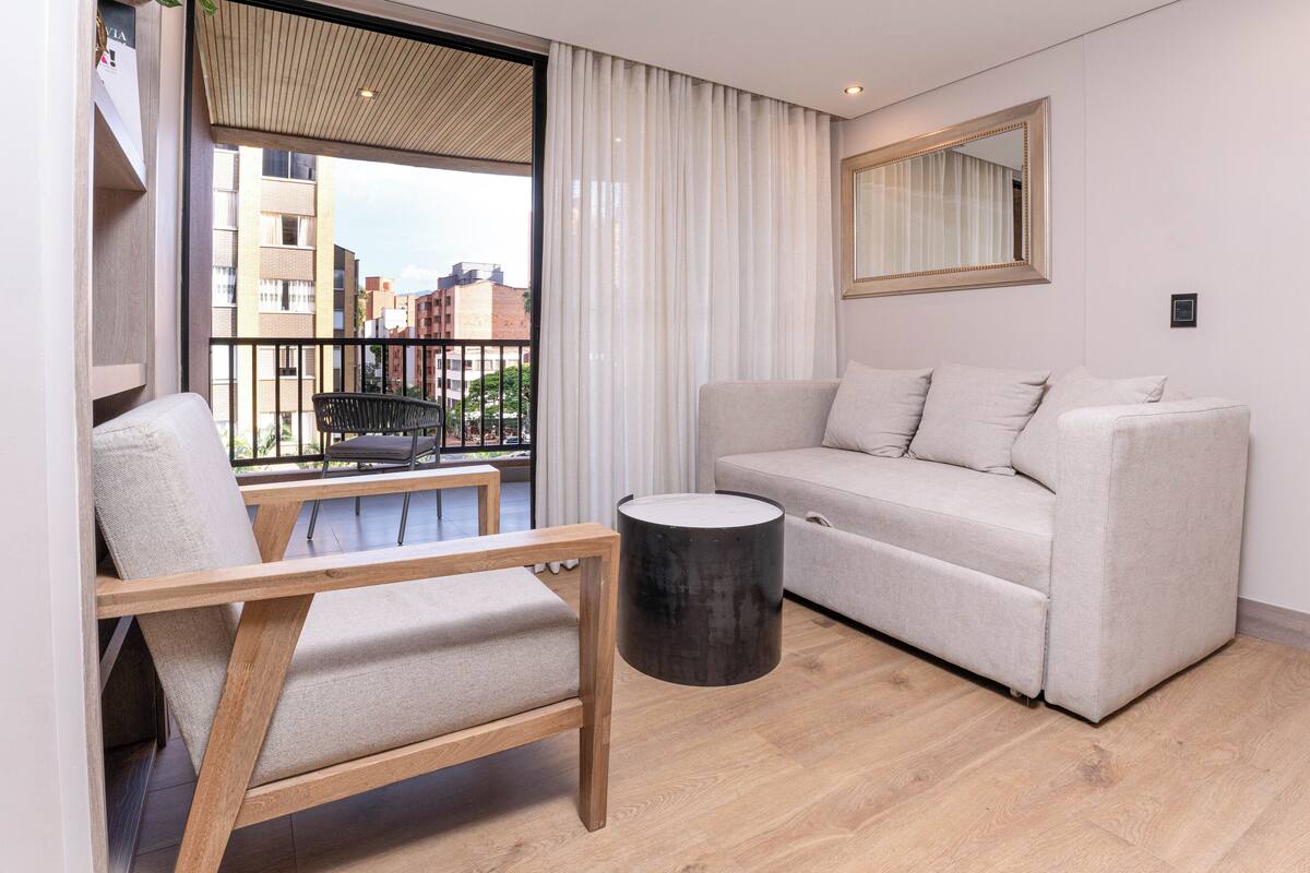 Un salon moderne avec un canapé, une chaise en bois, une petite table ronde et une vue sur les immeubles à travers une grande fenêtre.
