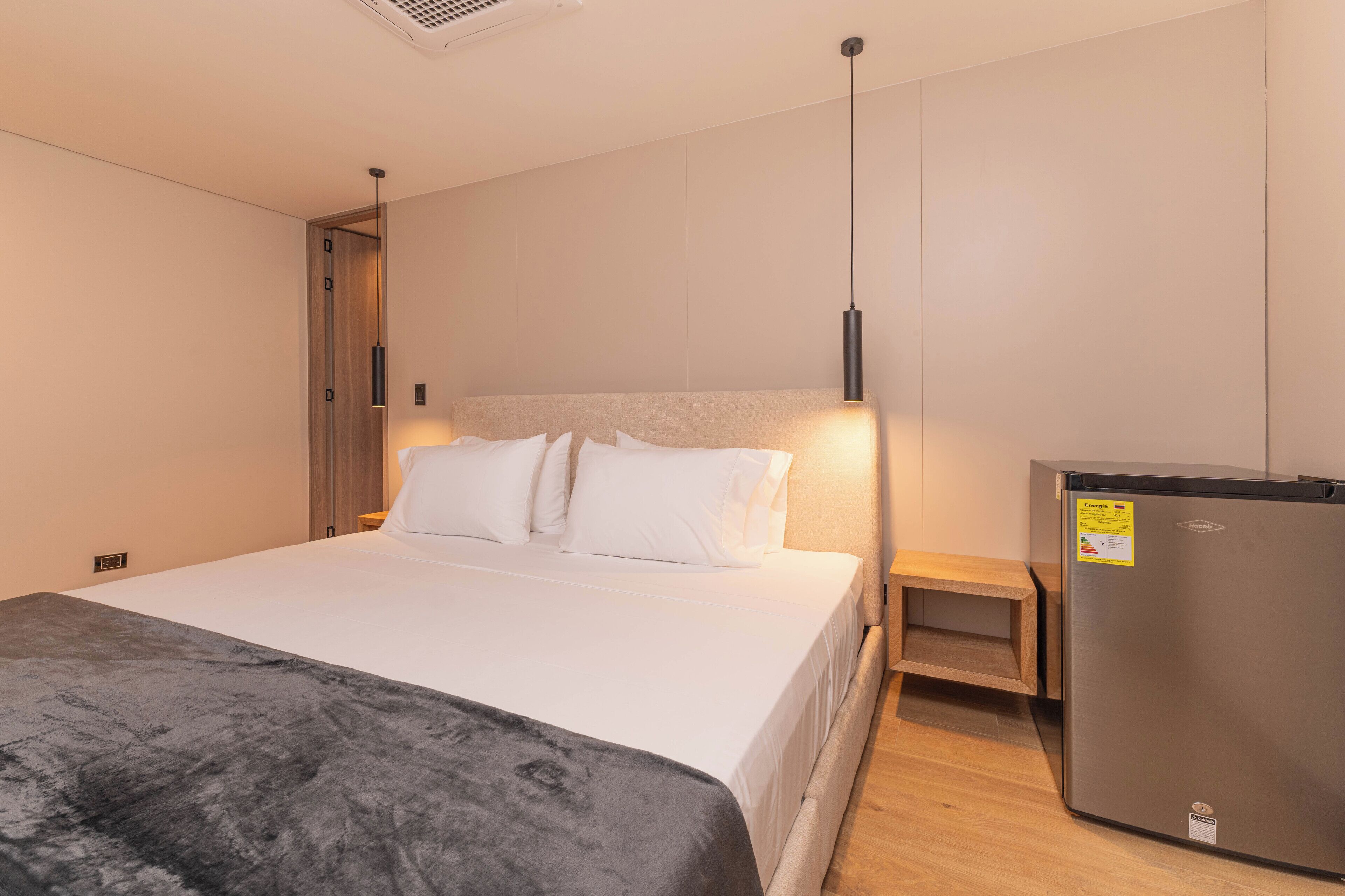 A hotel room with a bed, a nightstand, a wooden bench, and a refrigerator.