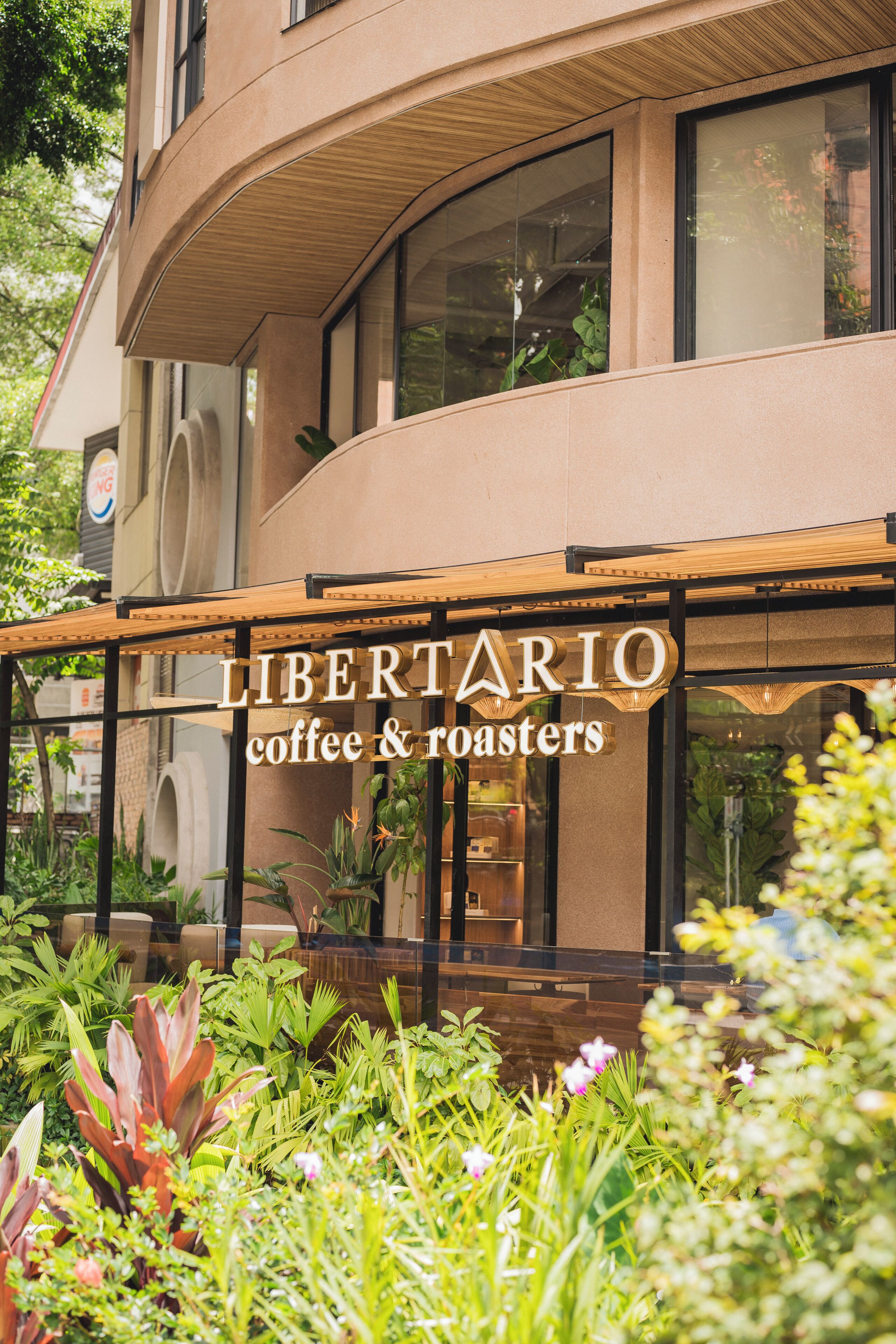 Ein modernes Gebäude mit einem Schild für 'LIBERTARIO coffee & roasters' und üppigem Grün davor.