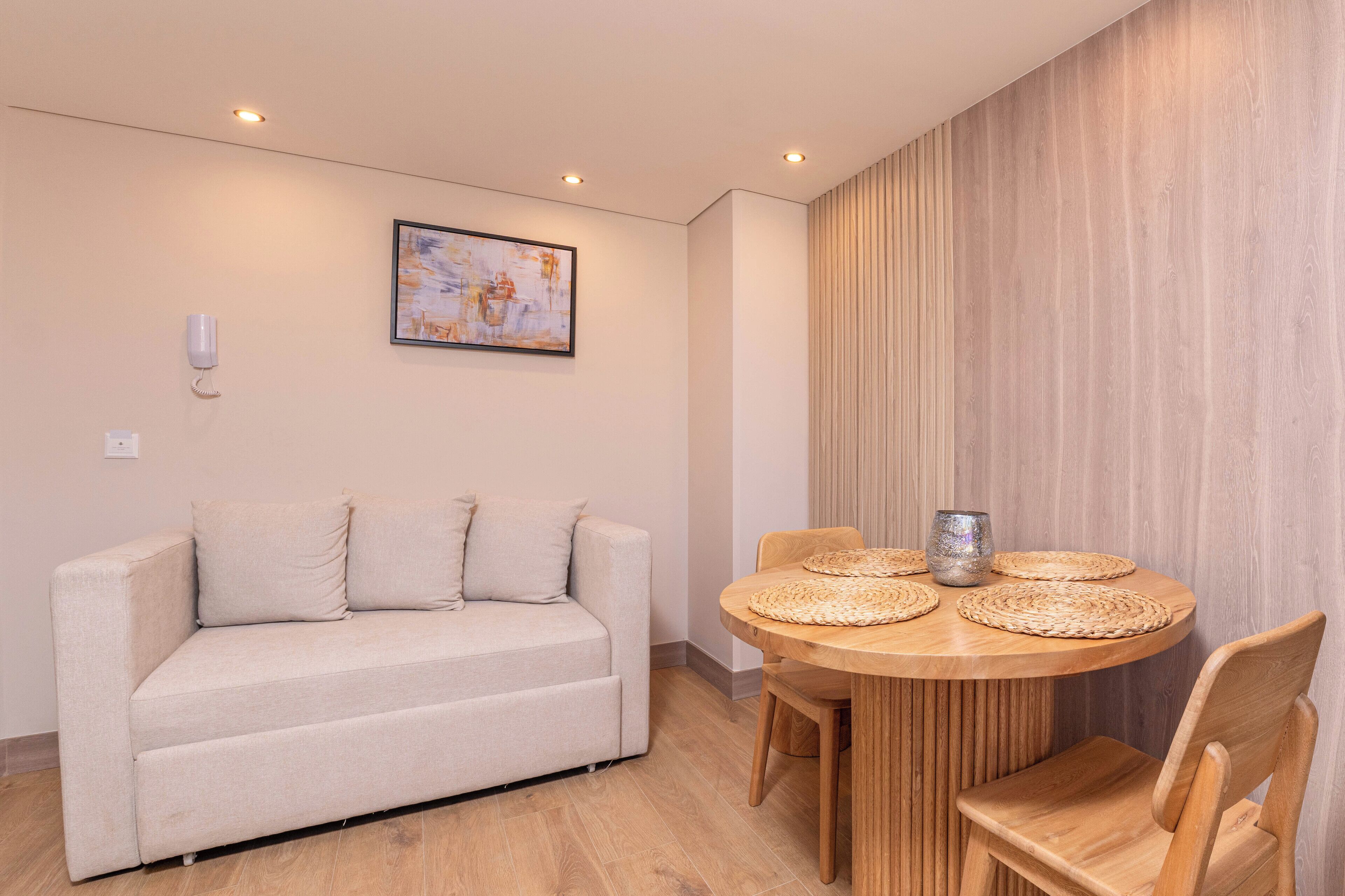 A living room with a beige sofa, a round wooden table with woven placemats, and a wall-mounted phone.