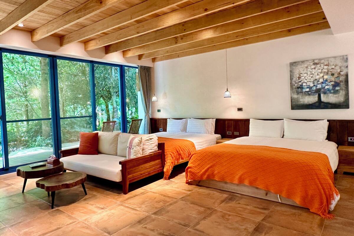 A bedroom with a large bed, two smaller beds, a sofa, and a small table. There are large windows with a view of trees.