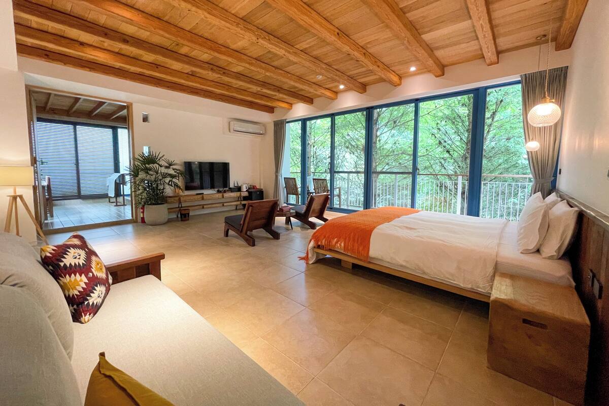 A spacious room with a large bed, a seating area with a sofa and chairs, a television, and a balcony with large windows.