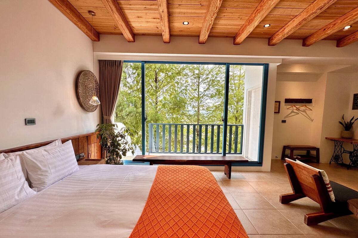 A bedroom with a bed, a bench, a table, and a view of trees through a large window.