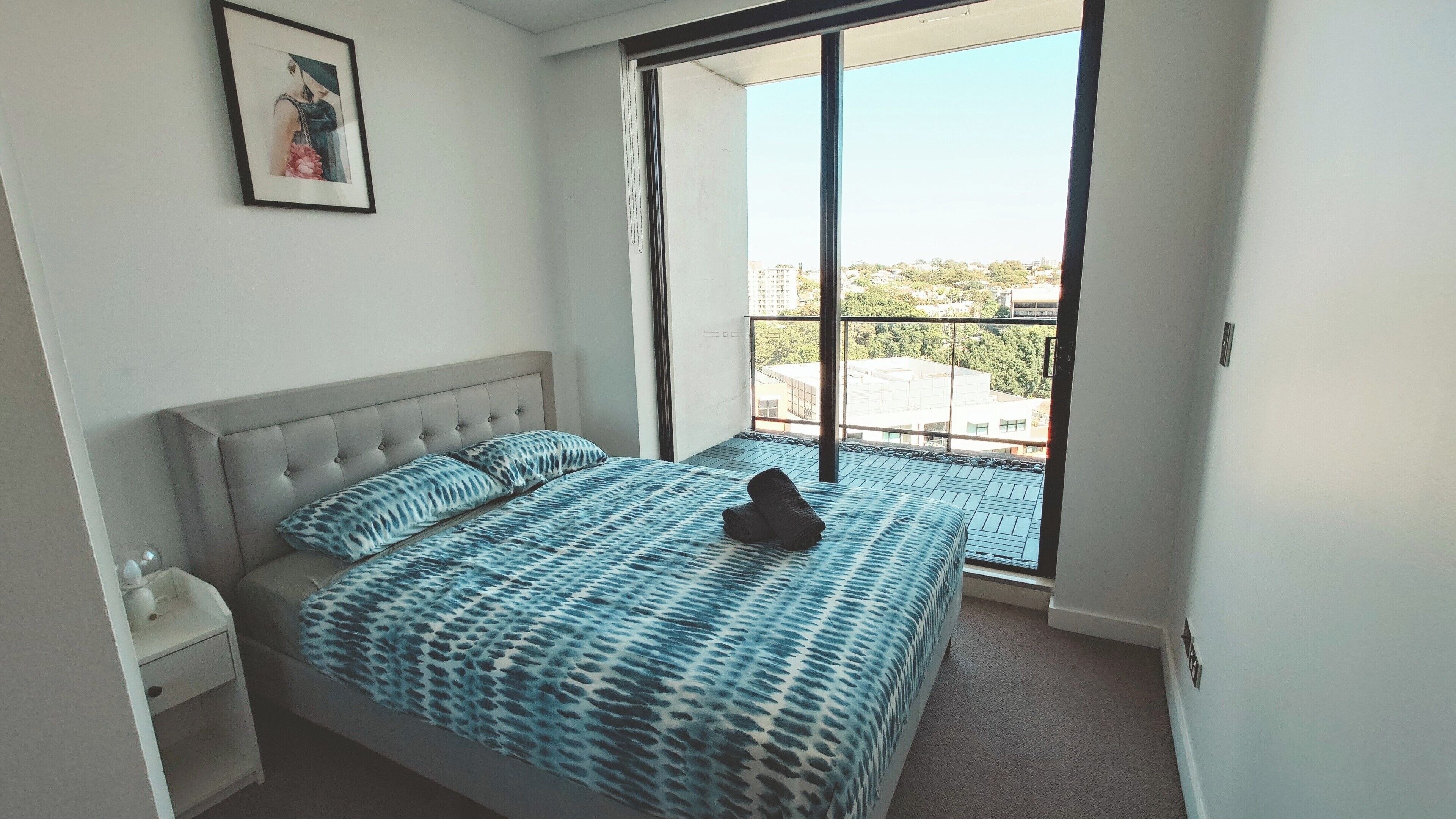 A bedroom with a large bed, a nightstand, a framed picture on the wall, and a view of the city through a large window.