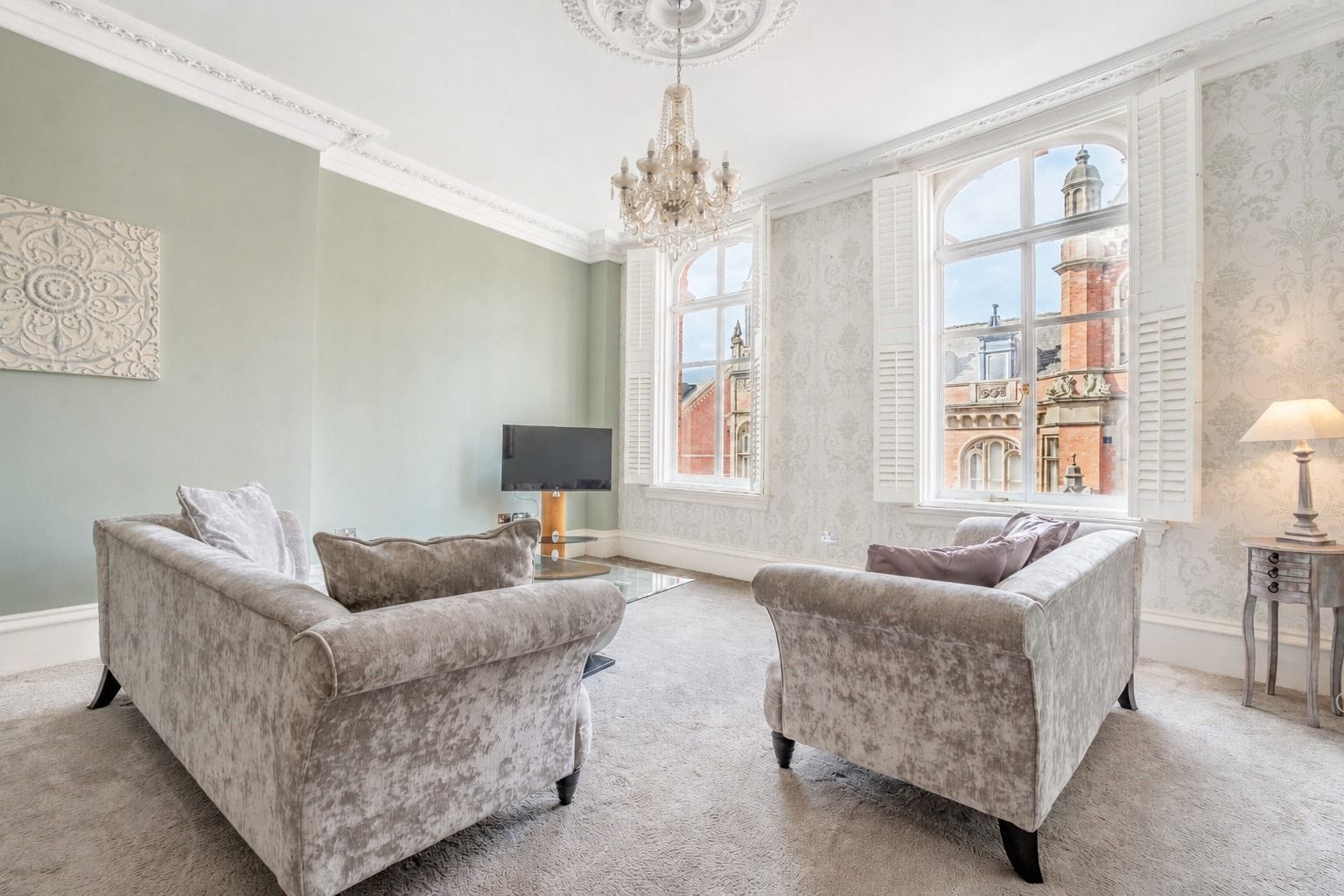 A spacious living room with two grey armchairs, a chandelier, and a large window with a view of a historic building.
