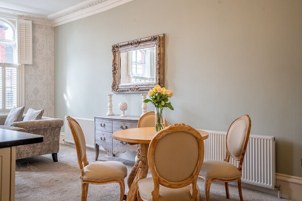Un coin repas comprenant une table ronde, quatre chaises rembourrées, un vase avec des fleurs, un miroir décoratif et un radiateur.