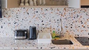 An open kitchen with a countertop, a sink, a coffee maker, a toaster, and a cabinet with dishes and glasses. - 1BR Condo Pool 5min Walk to Beach C131 (Choeng Thale)