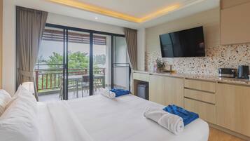 Une chambre d’hôtel avec un grand lit, un balcon avec une table et des chaises, un coin cuisine avec un téléviseur et une vue sur la verdure environnante.