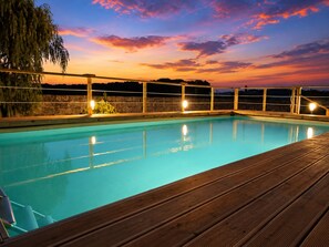 A swimming pool with a wooden deck, surrounded by a stone wall, and a sunset view.