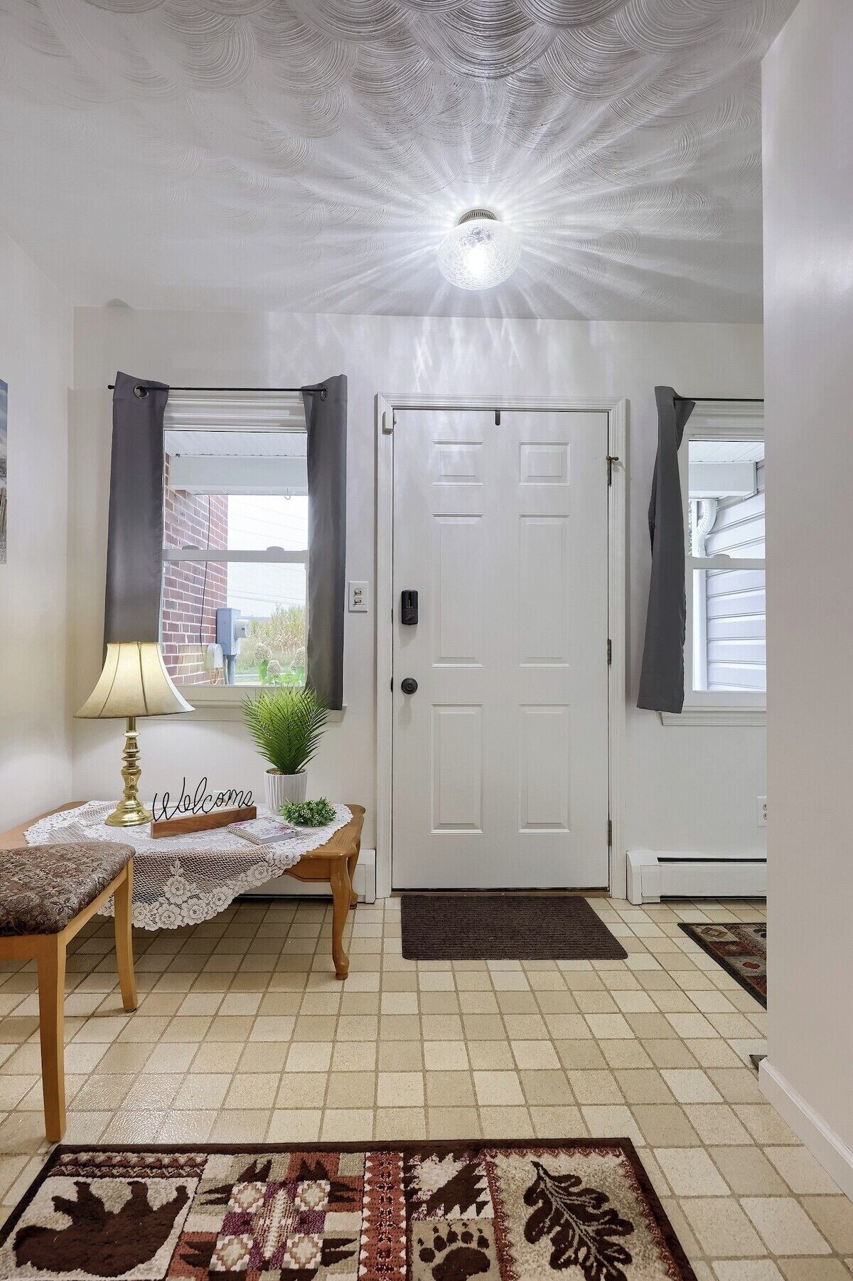 A room with a white door, a lamp, a bench, a welcome sign, and a patterned rug.