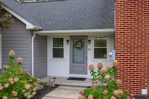 Uma casa com porta cinza, parede de tijolos e varanda com plantas de hortênsia.