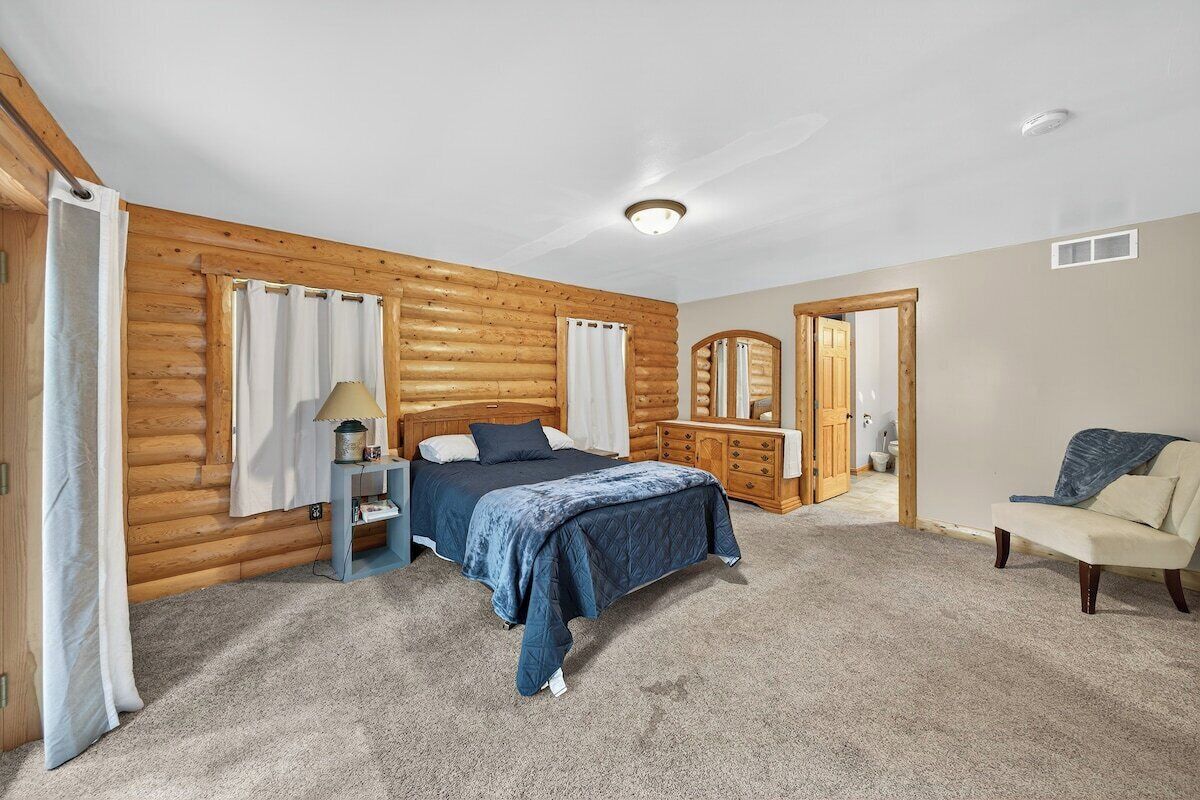 Quarto de cabana de toras com uma cama, mesa de cabeceira, penteadeira, espelho e uma cadeira.