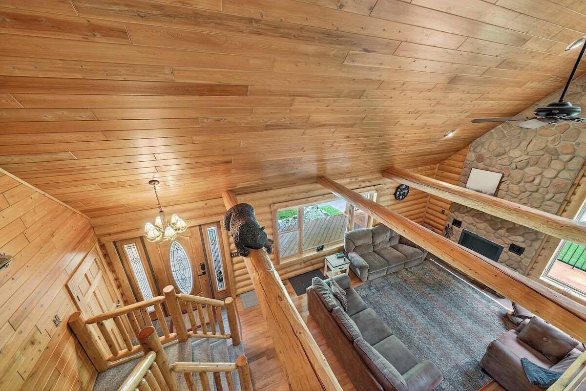 A log cabin interior with a wooden staircase, a living area with a sofa and a fireplace, and a stone wall.