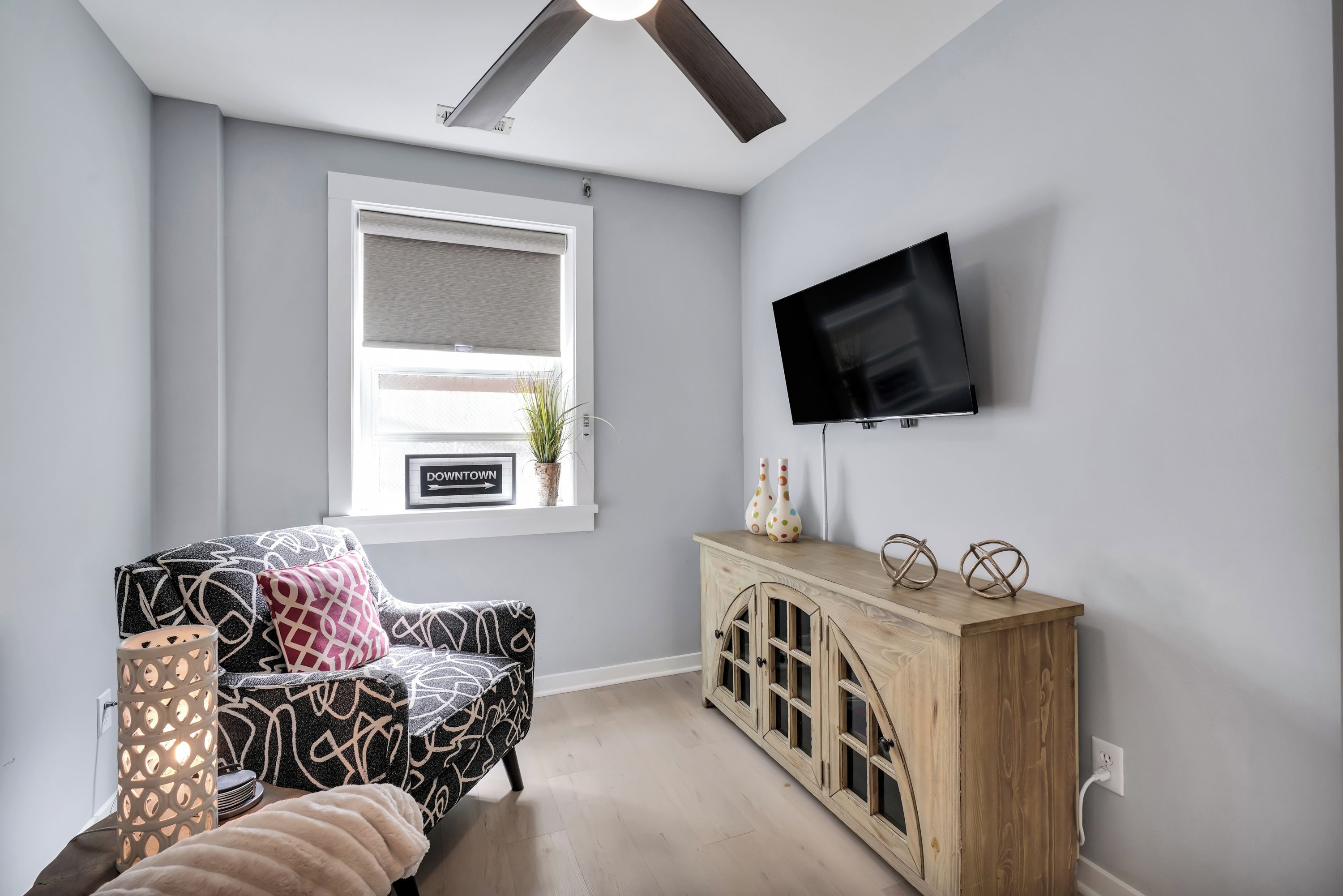 Una sala moderna con ventilador de techo, televisor de pantalla plana montado en la pared, ventana con vista al exterior, sofá estampado y mueble de madera con objetos decorativos.