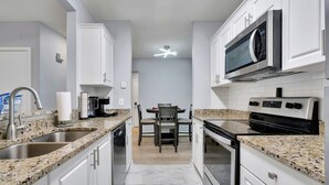 Una cocina moderna con encimeras de granito, armarios blancos, microondas integrado y un comedor con mesa y sillas.