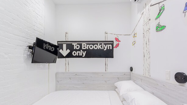 A room with a bed, a white brick wall, a sign pointing to Brooklyn, and a wall-mounted TV. - Compact Queen Retreat @ Times Square (New York)