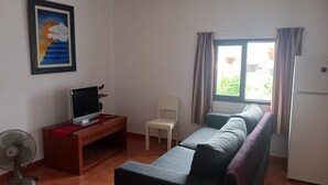 Ein kleines Wohnzimmer mit einer Couch, einem Fernseher, einem Sessel, einem Ventilator und einem Fenster mit Vorhängen.