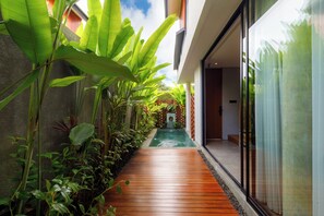 A narrow wooden pathway flanked by lush tropical plants leading to a modern building with large glass windows. - Damita E-2 by Dua Stay (Ubud)