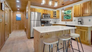 A kitchen with wooden cabinets, stainless steel appliances, a central island with stools, and a view of the outdoors through a window. - Bethel Hillside Chalet: Hot Tub + Fire Pit (Bethel)