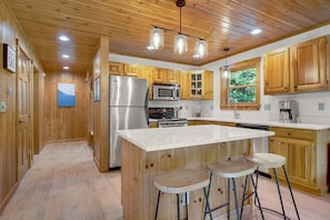 Una cocina con gabinetes de madera, electrodomésticos de acero inoxidable, una isla central con taburetes y vistas al exterior por una ventana.