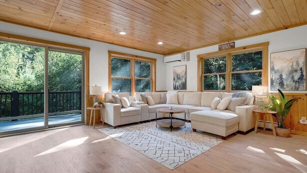 A spacious living room with a large sectional sofa, a coffee table, and a view of a forest through large windows. - Bethel Hillside Chalet: Hot Tub + Fire Pit (Bethel)