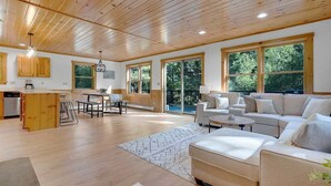 A spacious living area with a wooden ceiling, large windows, a kitchen with wooden cabinets, and a seating area with a sofa and coffee table. - Bethel Hillside Chalet: Hot Tub + Fire Pit (Bethel)