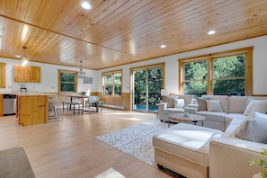 Amplia sala de estar con techo de madera, ventanas grandes, cocina con armarios de madera y un área de descanso con sofá y mesa de centro.
