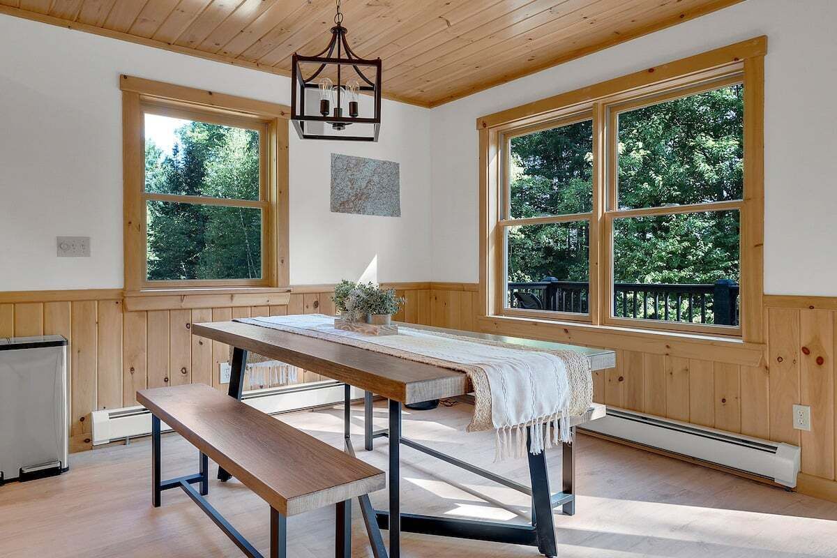 A room with a wooden bench, a table with a white cloth, and large windows with a view of trees.