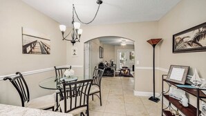 A dining area with a round glass table, black chairs, and a chandelier. There is a bookshelf with books and a framed picture on the wall. - What is missing from this Oceanside vacation  paradise?“YOU” (Cape Canaveral)