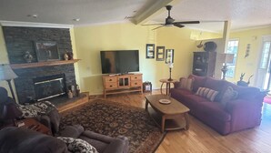 Un salon spacieux avec une cheminée, un grand téléviseur, un canapé, une table basse et un ventilateur de plafond.