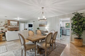 A dining area with a wooden table and chairs, a chandelier, and a view into the kitchen. - The Village Grove House | Renovated | Walk to Pier & Beach (st simons island)
