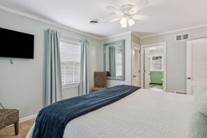 A bedroom with a bed, a ceiling fan, a television, and a bathroom visible through an open door. - The Village Grove House | Renovated | Walk to Pier & Beach (st simons island)