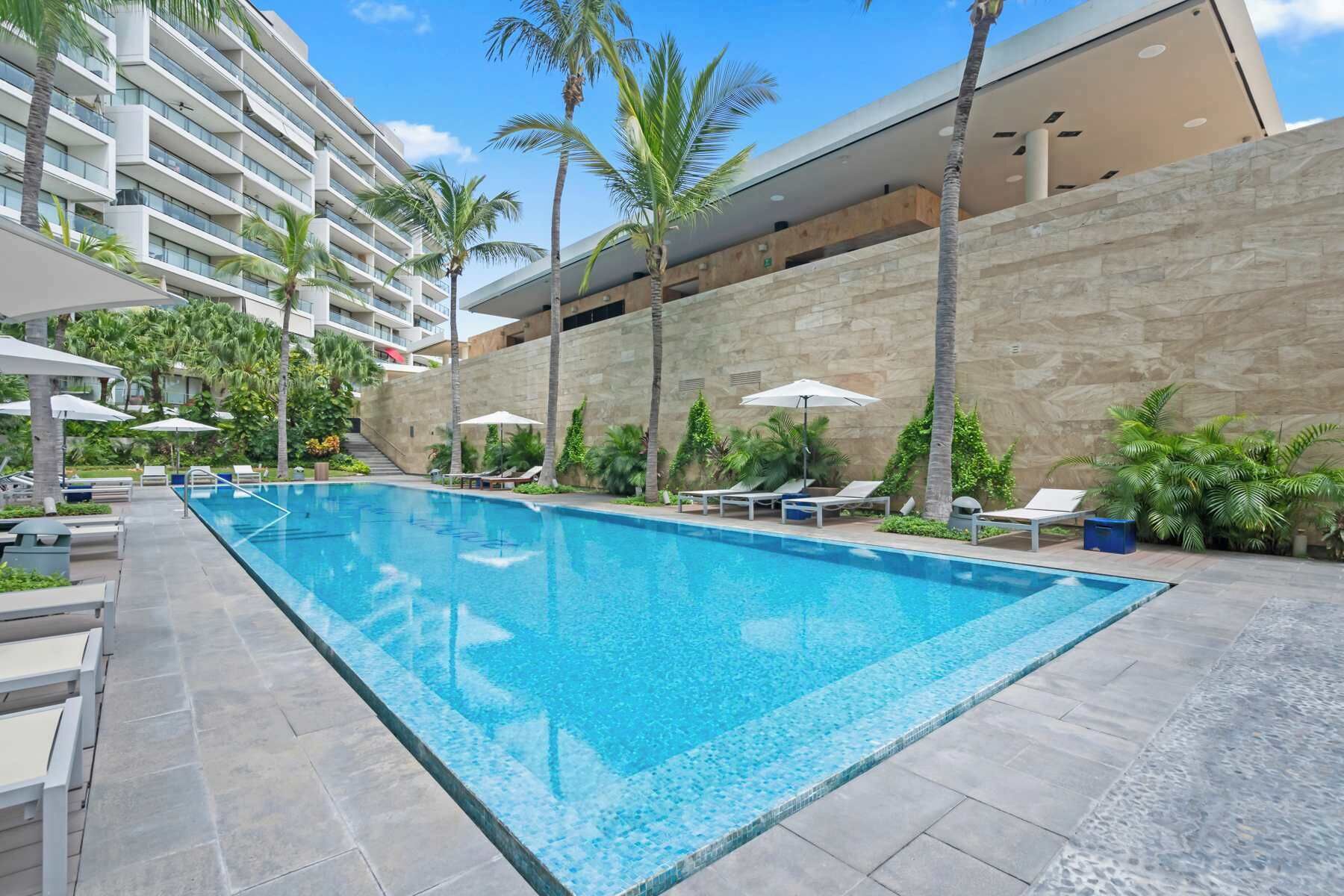 Una piscina con sillones de descanso y sombrillas, rodeada de palmeras y un muro de piedra.
