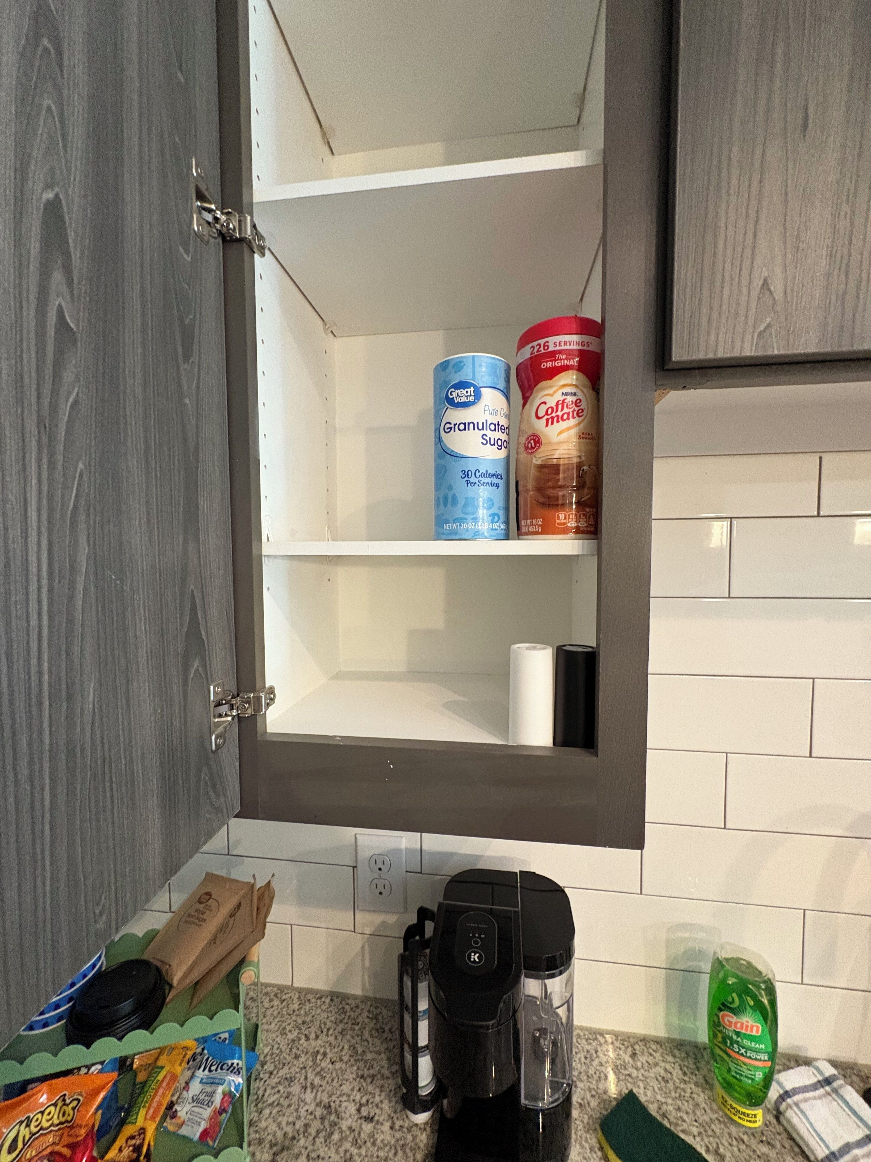 Dans la cuisine, on trouve un placard contenant un pot de sucre en poudre, une boîte de café et un sachet de crème pour café. Sur le comptoir, il y a une machine à café, une boîte de céréales et une bouteille de liquide vaisselle.