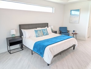 Una habitación de hotel moderna con una cama grande, dos mesitas de noche, una silla y una mesita con una lámpara.