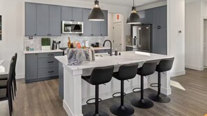 Una cocina moderna con gabinetes grises, una isla blanca con taburetes, electrodomésticos de acero inoxidable e iluminación colgante.