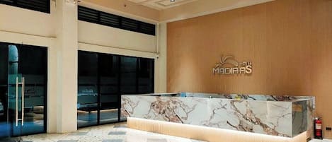 Zona de recepción de hotel con un mostrador de mármol, una pared de madera con el nombre del hotel y un piso de baldosas.